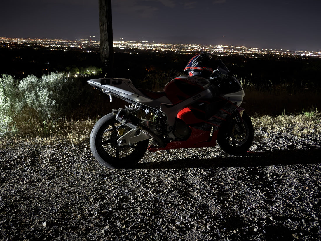 A sport bike with its kickstand down on a gravel overlook, helmet resting on the seat, city lights spread out below at dusk