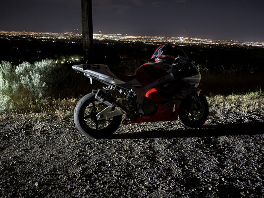 A sport bike with its kickstand down on a gravel overlook, helmet resting on the seat, city lights spread out below at dusk